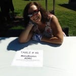 Person smiles for a photo at their table. Sign on table reads "Table #46 MetroBoston ADRC".