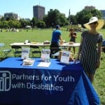 A person in a striped dress and wide brimmed hat stands next to a table with a blue tablecloth. Tablecloth reads "Partners for Youth with Disabilities."