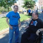 A person in a black ADA 25 t-shirt speaks with a person in a blue NILP t-shirt.