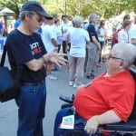 A person wearing a black t-shirt speaks to a person in a red polo seated in a wheelchair.