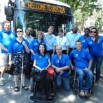 A group of people in blue polos stand for a group photo in front of an MBTA bus. The top of the bus reads "Welcome to Boston."