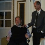 A BCIL staff member in a dark gray suit holds the microphone for a speaker in a wheelchair.