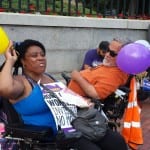 A person in a wheelchair cheers and holds a yellow balloon in their hand. Behind them is another person in a wheelchair with a purple balloon.