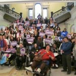 A large group of people, some in wheelchairs, have group photo on main staircase. People hold signs which say United for Home Care.