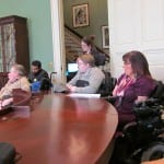 People seated in wheelchairs listen to a speaker.