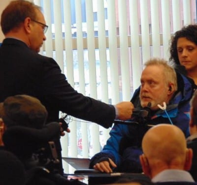 A person in a dark suit jacket, blue shirt and yellow tie speaks to a person in a wheelchair.