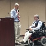 Person at podium holds microphone and speaks to person in wheelchair.