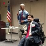 Person in blue shirt holds microphone while standing next to man in wheelchair.