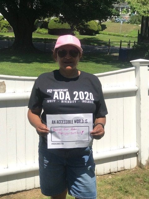 A person wearing a black ADA 30 t-shirt holds a sign that read AN ACCESSIBLE WORLD IS BETTER FREE PUBLIC TRANSPORTATION!