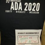 A black ADA 30 t-shirt and a sign that reads DISABILITY DISCRIMINATION IS AN ACT THAT NEEDS TO STOP NOW.