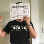 A person wearing a black ADA 30 t-shirt holds a sign that reads DISABILITY DISCRIMINATION IS WRONG in front of their face.