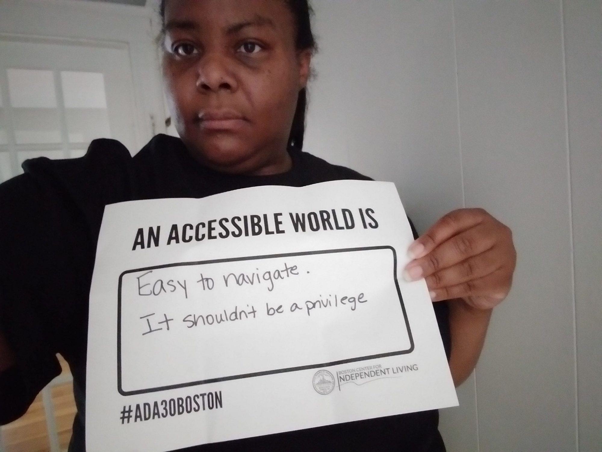 A person wearing a black ADA 30 t-shirt holds a sign that reads AN ACCESSIBLE WORLD IS EASY TO NAVIGATE, IT SHOULDN'T BE A PRIVILEGE.