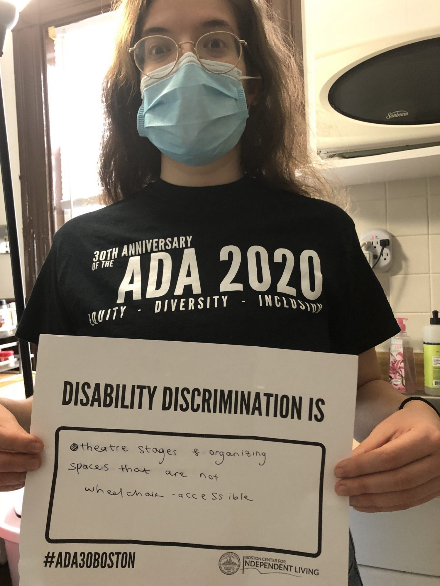 A person wearing a black ADA 30 t-shirt and blue face mask holds up a sign saying DISABILITY DISCRIMINATION THEATRE STAGES & ORGANIZING SPACES THAT ARE NOT WHEELCHAIR ACCESSIBLE.