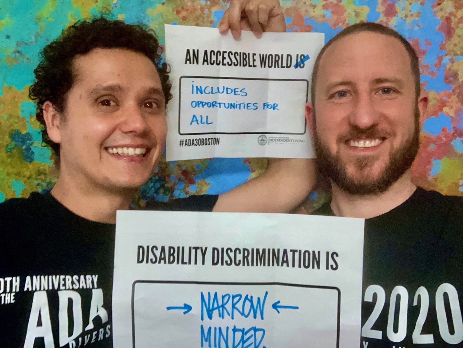 Two people wearing black ADA 30 t-shirts hold signs that read AN ACCESSIBLE WORLD INCLUDES OPPORTUNITIES FOR ALL and DISABILITY DISCRIMINATION IS NARROW MINDED.