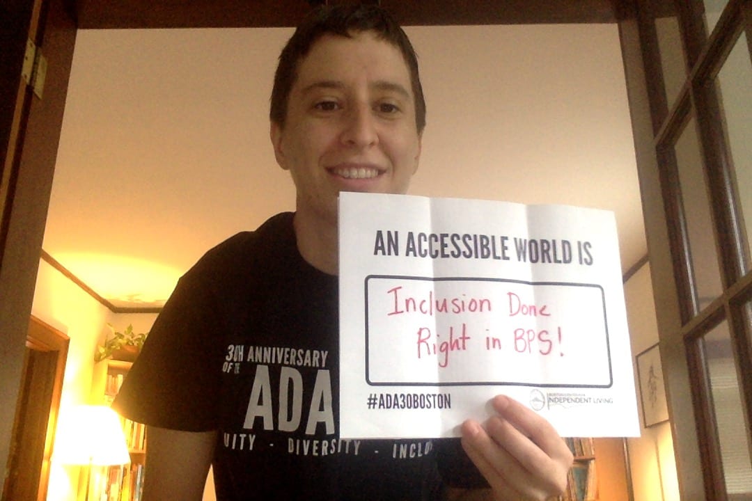 A person wearing a black ADA 30 t-shirt holds a sign that read AN ACCESSIBLE WORLD IS INCLUSION DONE RIGHT IN BPS!