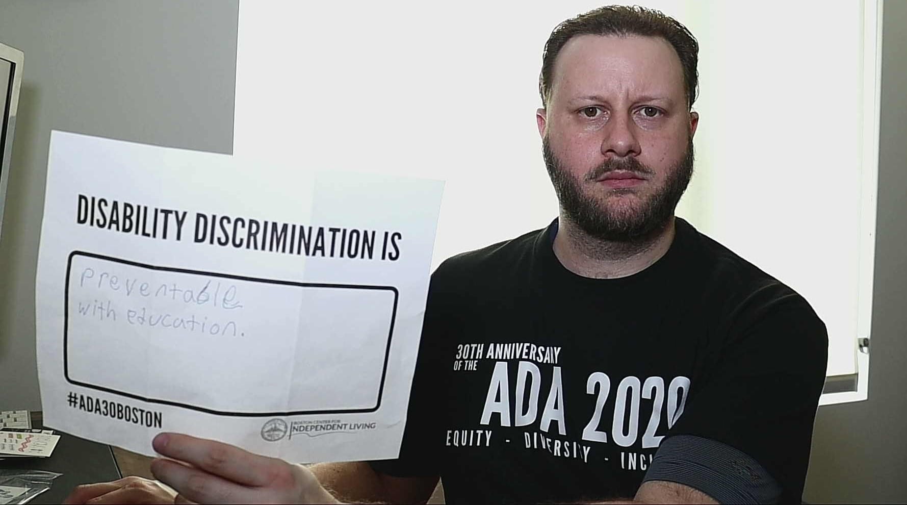 A person in a black ADA 30 t-shirt holds up a sign saying DISABILITY DISCRIMINATION IS PREVENTABLE WITH EDUCATION.