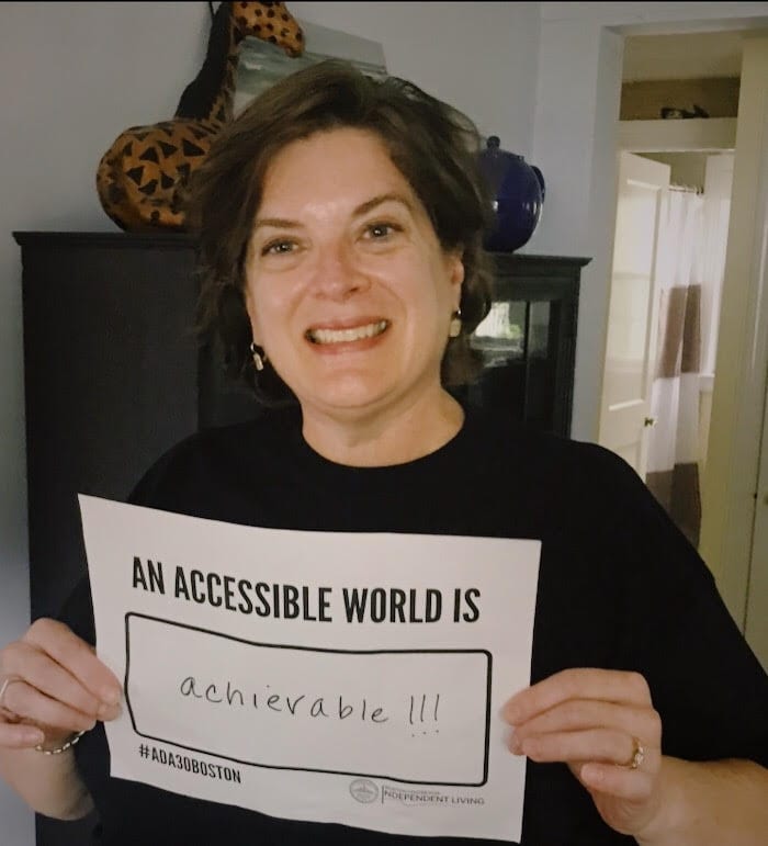 A person in a black ADA 30 t-shirt holds up a sign saying AN ACCESSIBLE WORLD IS ACHIEVABLE!!!