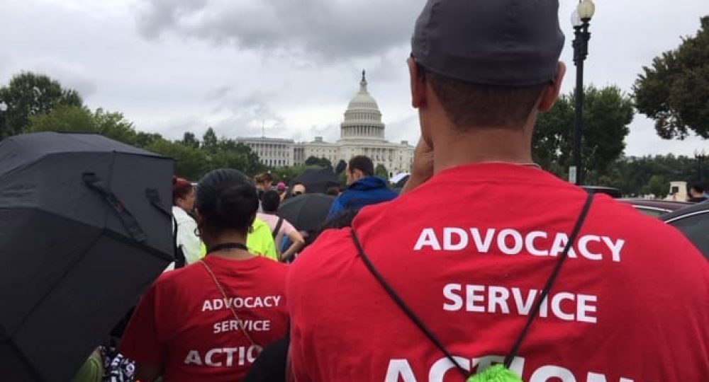 A large crowd of people gather outside the U.S. Capitol. In the photo's foreground, two people wear red t-shirts that say ADVOCACY SERVICE ACTION! on the back. A person holds a black umbrella off to the left.